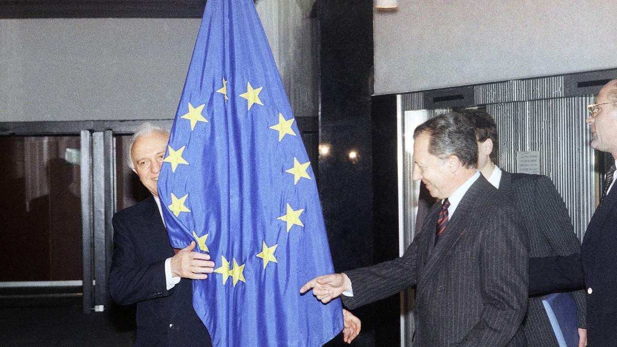 El ministro soviético de Asuntos Exteriores, Eduard Shevardnadze, se asoma por detrás de la bandera de la Unión Europea, mientras Jacques Delors señala el lugar donde debería estar de pie; imagen de 1989