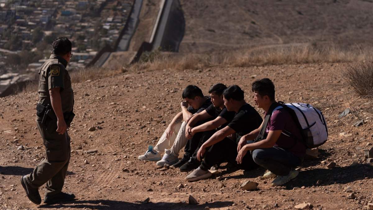 Un agente de la Patrulla Fronteriza pasa junto a cuatro hombres detenidos después de cruzar ilegalmente la frontera México-EU, en San Diego