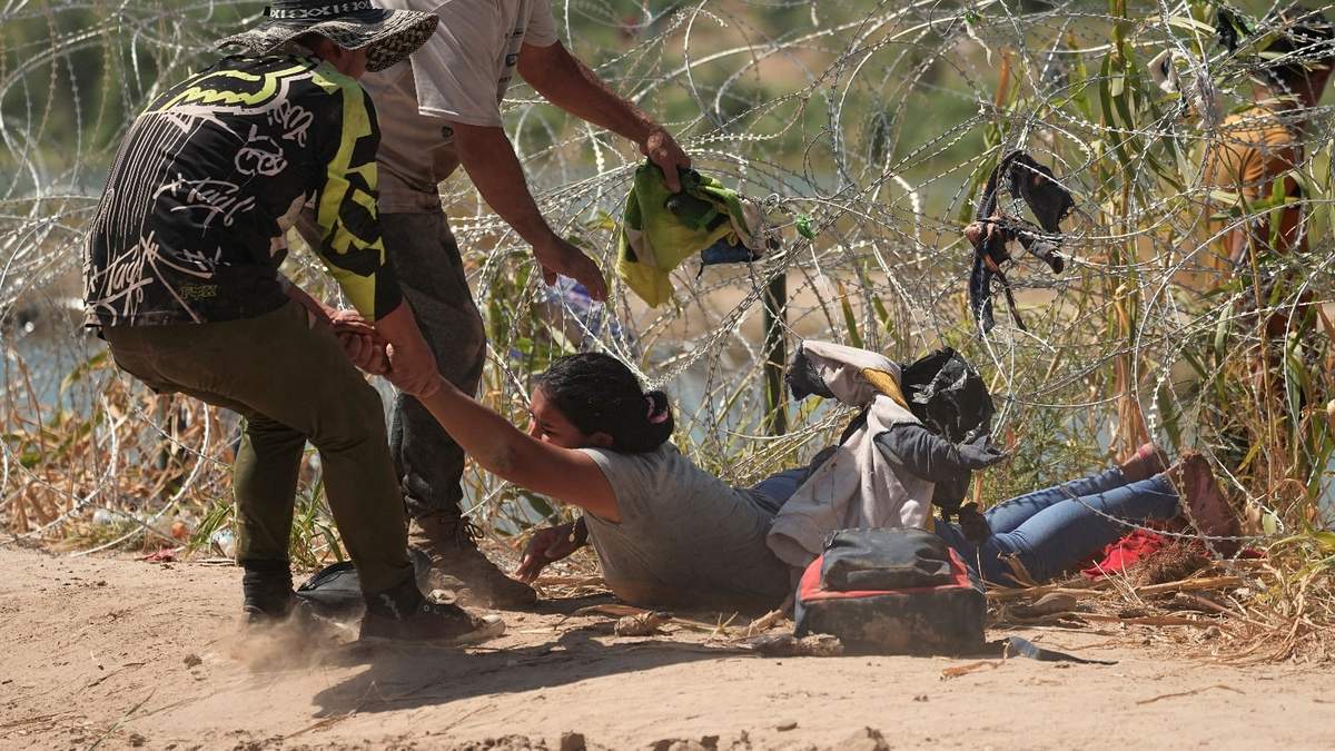 Una inmigrante que cruzó a Estados Unidos desde México es auxiliada a pasar debajo de alambre de púas en una de las márgenes del río Bravo, el jueves 21 de septiembre de 2023, en Eagle Pass, Texas