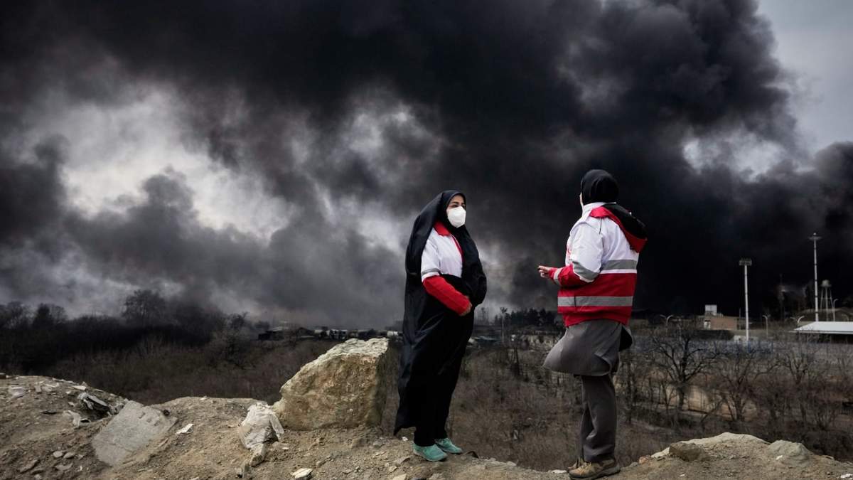 Dos mujeres de la Sociedad de la Luna Roja se ven de pie con una densa humareda provocada por un ataque el sábado por la noche de la campaña estadounidense e israelí contra una instalación petrolera, en Teherán, Irán, el domino 8 de marzo de 2026
