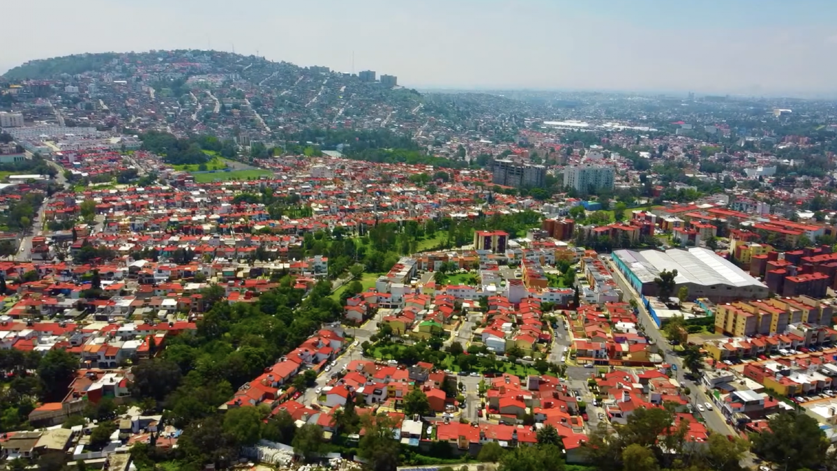 Paisaje urbano de Atizapán de Zaragoza bajo un cielo nublado, con calles húmedas debido a las precipitaciones ligeras típicas de la temporada. Los habitantes caminan con paraguas y ropa abrigadora, reflejando el ambiente templado pero inestable que predomina en esta región del Estado de México durante la jornada dominical.