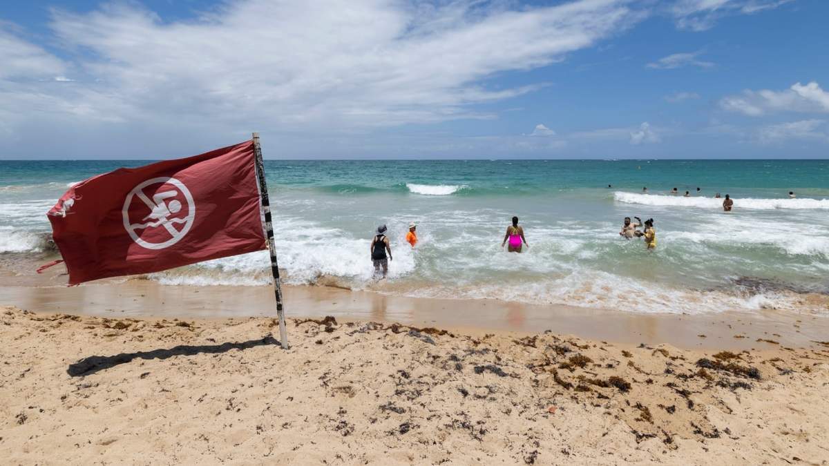Una bandera de advertencia ondea al viento en una playa mientras la gente se baña en Condado, Puerto Rico, mientras el huracán Erin se acerca