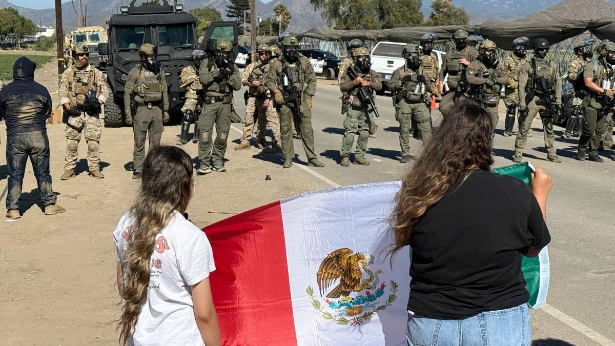 Manifestantes sostienen una bandera frente a agentes federales que bloquean un camino durante una redada de inmigración en Camarillo, California