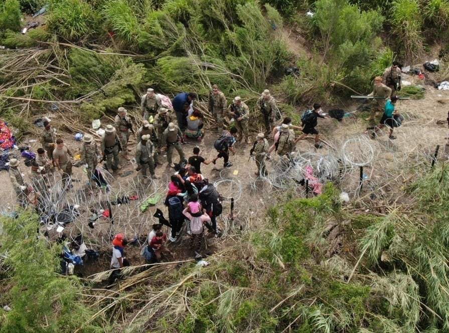 Migrantes se paran en la orilla estadounidense del río Bravo, donde la Guardia Nacional de Texas se para detrás de alambre de púas, visto desde Matamoros, México. Las restricciones de asilo de EU relacionadas con la pandemia, conocidas como Título 42, expirarán este 11 de mayo