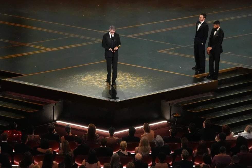 Paul Thomas Anderson recibe el premio al mejor guion adaptado por “One Battle After Another” durante la ceremonia de los Óscar el domingo 15 de marzo de 2026 en el Dolby Theatre de Los Ángeles. Chris Evans, segundo por la derecha, y Robert Downey Jr. observan desde la derecha.