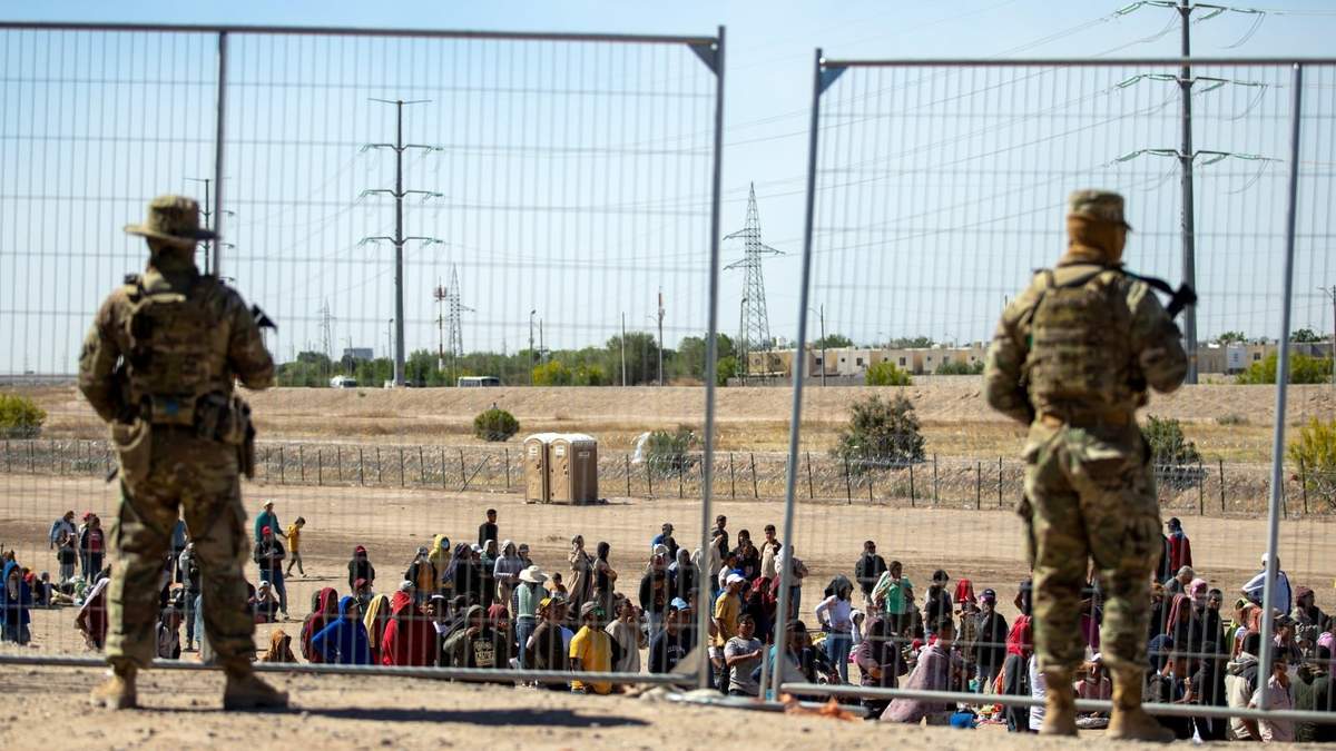 Migrantes esperan junto a una valla fronteriza bajo la vigilancia de la Guardia Nacional de Texas