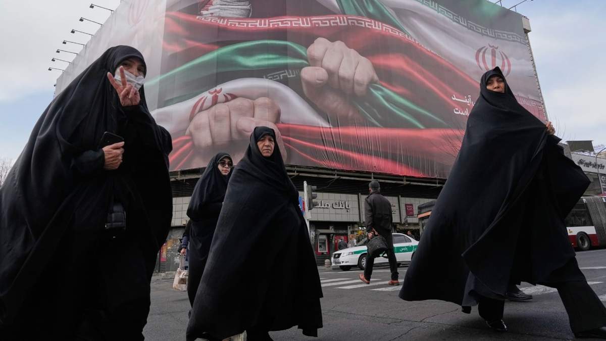 Las mujeres cruzan una calle bajo una enorme pancarta que muestra manos que sostienen firmemente banderas iraníes como señal de patriotismo, mientras una de ellas muestra el signo de la victoria, en Teherán