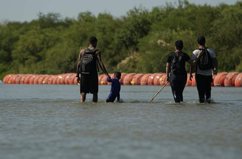 Migrantes en su ruta hacia EU caminan a lo largo de las boyas instaladas como barrera sobre el Río Bravo
