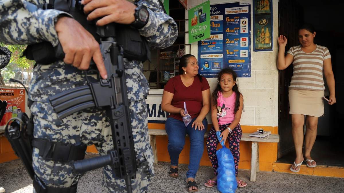 Varias personas sentadas en la entrada de una tienda custodiada por un militar en San Antonio Los Ranchos, El Salvador